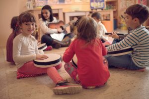 Happy little girl having a music class with her friends and teacher in a preschool and looking at the camera.
